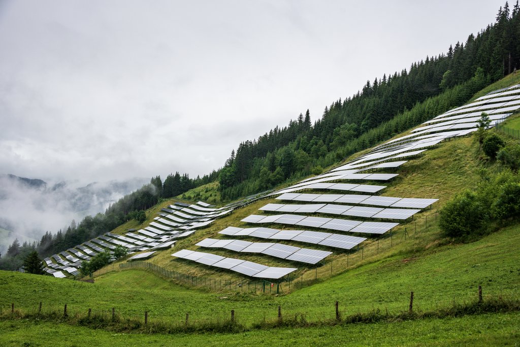 Solar, photovoltaic power station located on a mountain slope in the Alps. Foggy day, green pasture grass and blue skies and mountains create a fresh and ecological image.